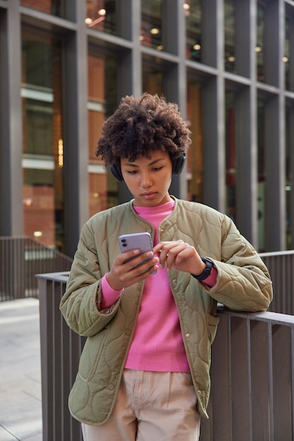 Pretty serious young woman with curly hair reads income message on modern smartphone stands against urban setting background listens music via headphones searches location via phone application