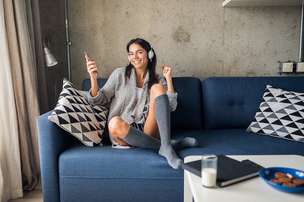 Pretty sexy smiling woman in casual outfit sitting in living room listening to music on headphones, having fun at home