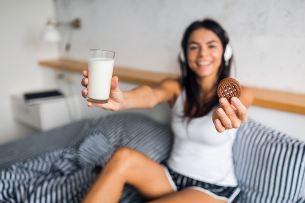Довольно сексуальная улыбающаяся женщина сидит в постели утром, слушает музыку в наушниках, завтракает, ест печенье и пьет молоко, здоровый образ жизни