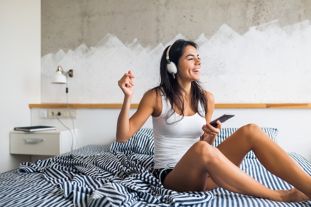 Donna sorridente abbastanza sexy che si siede a letto la mattina, ascoltando musica in cuffia, divertendosi