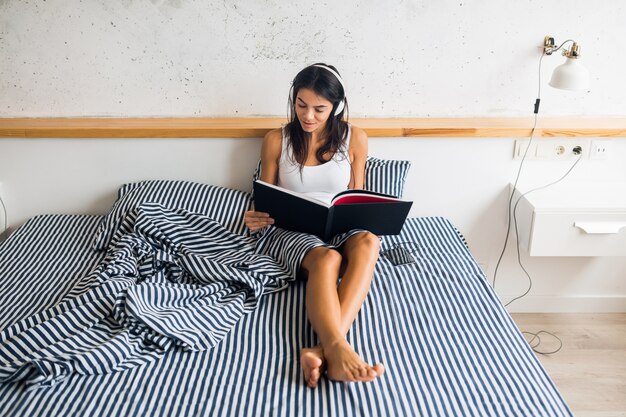 Pretty sexy smiling woman sitting in bed in morning, listening to music on headphones and reading book