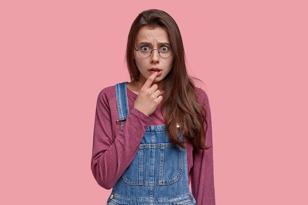 Pretty shocked frightened young woman keeps finger on lip, looks with frustration, has long dark hair, wears denim dungarees