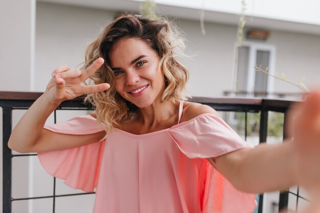 Pretty short-haired caucasian female model making selfie with peace sign. Glad tanned girl in pink dress taking picture of herself and smiling at balcony.