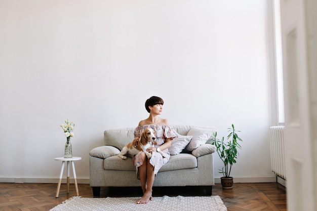 Pretty short-haired lady thoughtfully looking away while beagle dog resting on her knees