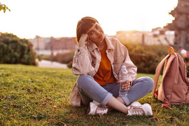 Pretty short-haired woman in jeans and pink jacket sits on grass during sunset