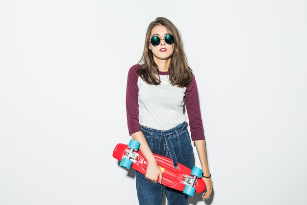 Pretty skater girl in casual clothes and black sunglasses holding red skateboard isolated on white wall