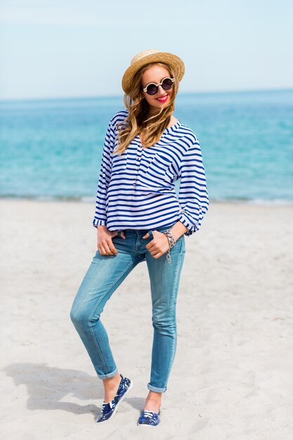 Pretty slim tan blonde stylish woman in straw hat and sunglasses, posing on the paradise tropical beach