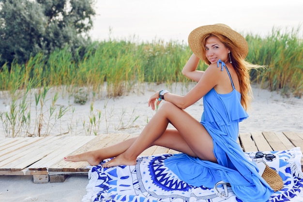 해변에서 놀라운 휴가 시간을 보내는 밀짚 모자에 긴 붉은 머리카락과 함께 꽤 슬림 여자. 파란 드레스를 입고. 세련된 커버에 앉아 있습니다.