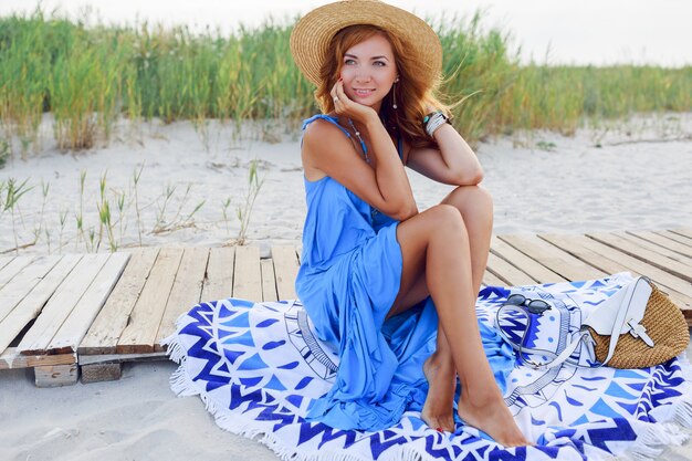 Pretty  slim woman with long red hairs in straw hat spending amazing holiday time on the beach. Wearing blue dress. Sitting on stylish cover.