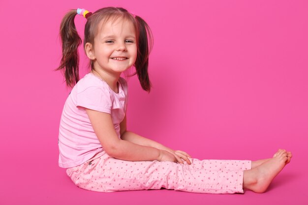 Pretty small kid with two pony tails and many colourful scrunchies sits on floor and glad to be photographed in photo studio. Adorable child smiles. Children and childhood concept.