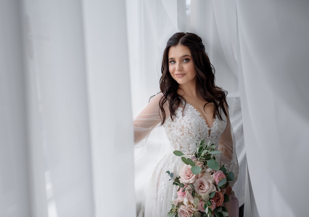 Pretty smiled brunette bride is holding tender wedding bouquet near the window and looking straight