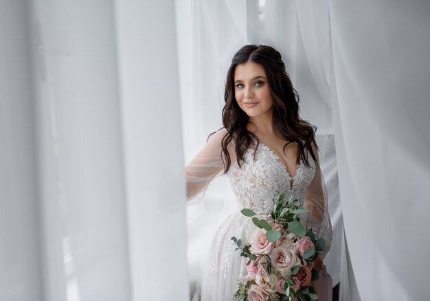 Pretty smiled brunette bride is holding tender wedding bouquet near the window and looking straight