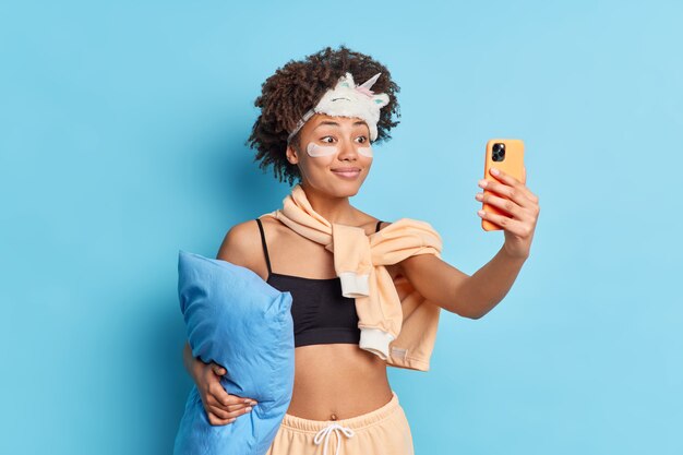 Pretty smiling African American woman in nightwear sleepmask holds pillow takes selfie via mobile phone applies collagen patches