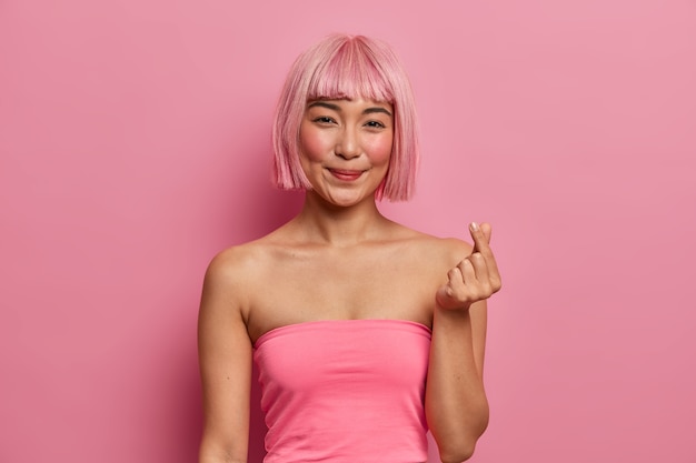 Pretty smiling Asian lady with bob hairstyle, expresses love, makes like gesture with fingers, wears rosy top