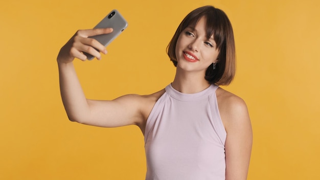 Pretty smiling brunette girl with bob hair and red lips making selfie on smartphone for social network isolated on yellow background