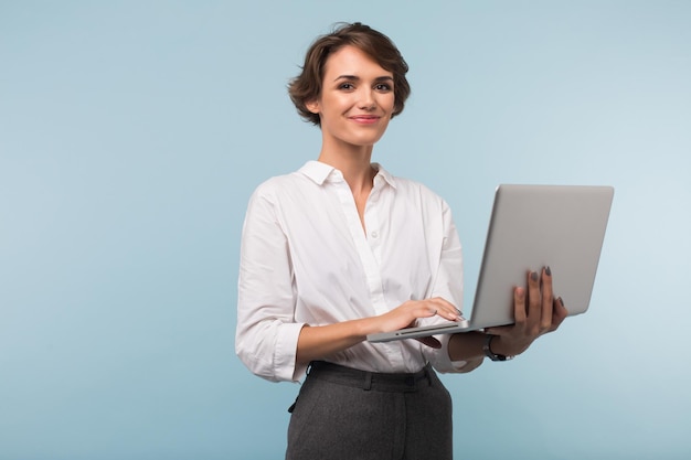 Pretty smiling businesswoman with dark short hair in white shirt holding laptop in hands while happily looking in camera over blue background