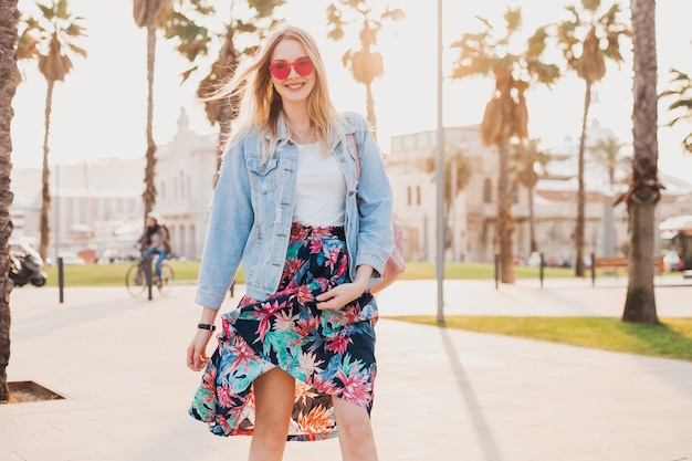 Довольно улыбающаяся флиртующая романтическая женщина гуляет по городской улице в стильной юбке с принтом и джинсовой куртке oversize в розовых солнцезащитных очках