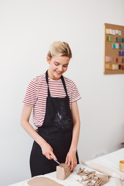검은 앞치마와 줄무늬 티셔츠를 입은 예쁜 미소녀가 현대 도자기 스튜디오에서 즐겁게 일하고 있다