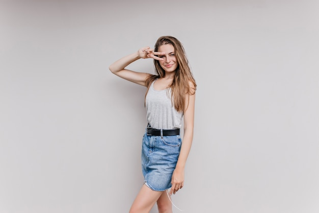 Pretty smiling girl in denim skirt posing with peace sign. Indoor portrait of wonderful caucasian  lady having fun.