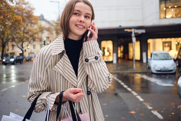 Симпатичная улыбающаяся девушка счастливо разговаривает по смартфону, гуляя по уютной городской улице с сумками для покупок