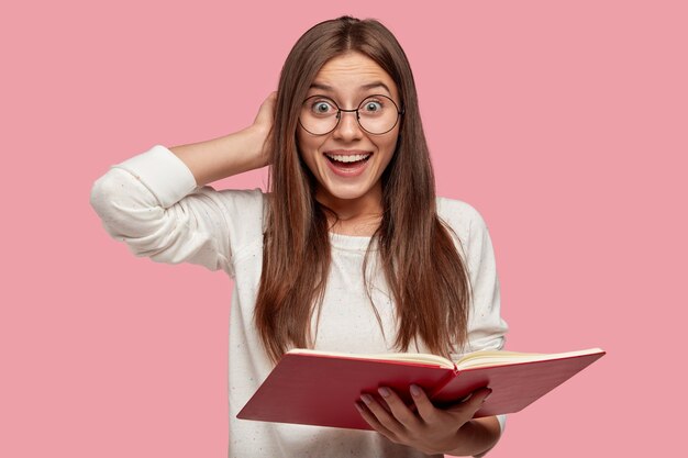 Pretty smiling girl has joyful expression, keeps hand behind head, carries red textbook, smiles broadly as reads necessary information, isolated over pink wall, wears round spectacles.