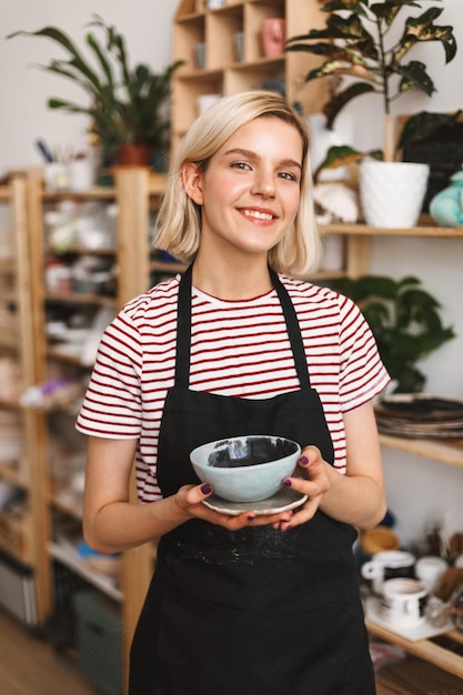 Бесплатное фото Симпатичная улыбающаяся девушка в черном фартуке и полосатой футболке, держащая в руках тарелку и миску ручной работы, счастливо смотрит в камеру в гончарной мастерской