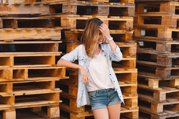 Ragazza sorridente graziosa, jeans da portare, shorts e camicia che propongono sulla priorità bassa di legno