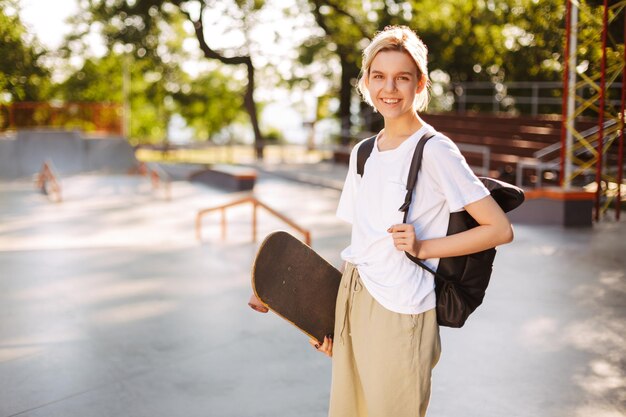 Pretty smiling girl with backpack and skateboard in hand joyfully looking in camera with big modern skatepark on background