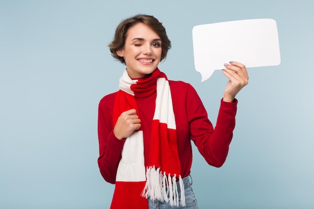 Pretty smiling girl with dark short hair in red sweater and scarf happily winking while looking in camera with white paper shape of message in hand over blue background