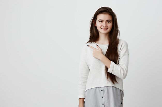 Pretty smiling joyfully female with dark long hair, pointing with her index finger away, showing copy space