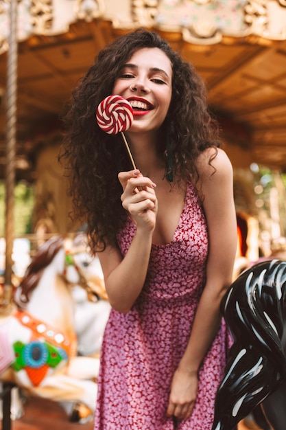 手にキャンディーポップキャンディーを手に立って、背景にカルーセルと遊園地で時間を過ごしている間、カメラで幸せに見てドレスを着て暗い巻き毛のかわいい笑顔の女性