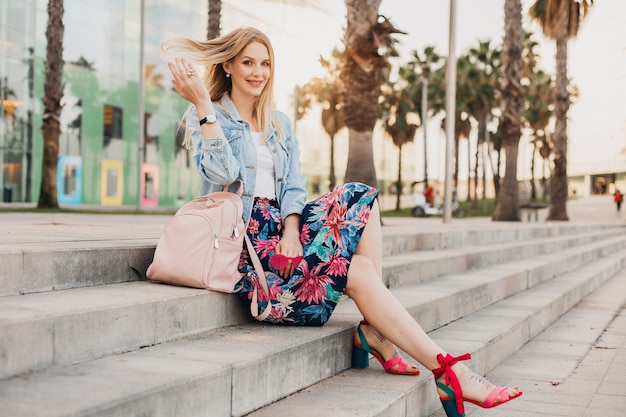 Pretty smiling woman sitting on stairs in city street in stylish printed skirt and denim oversize jacket with leather backpack, summer style trend