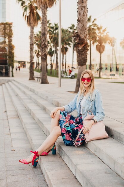 Pretty smiling woman sitting on stairs in city street in stylish printed skirt and denim oversize jacket with leather backpack wearing pink sunglasses, summer style trend