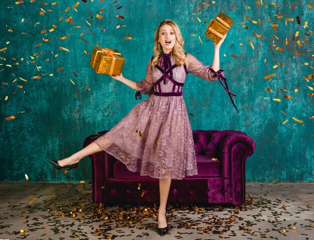 Pretty smiling woman in stylish violet evening dress against velvet sofa with gifts