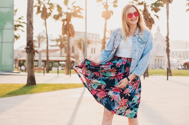 Pretty smiling woman walking in city street in stylish printed skirt and denim oversize jacket wearing pink sunglasses, summer style trend