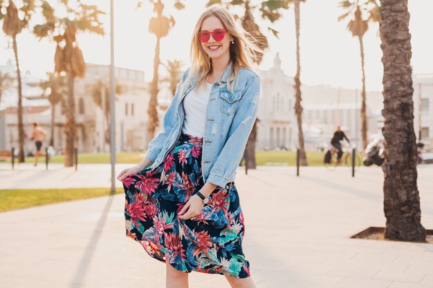 Довольно улыбающаяся женщина гуляет по городской улице в стильной юбке с принтом и джинсовой куртке oversize в розовых солнцезащитных очках, летний тренд