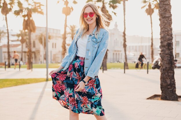 Pretty smiling woman walking in city street in stylish printed skirt and denim oversize jacket wearing pink sunglasses, summer style trend