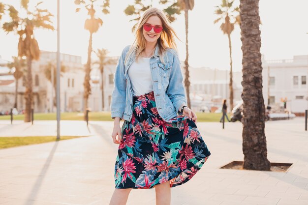 Pretty smiling woman walking in city street in stylish printed skirt and denim oversize jacket wearing pink sunglasses, summer style trend