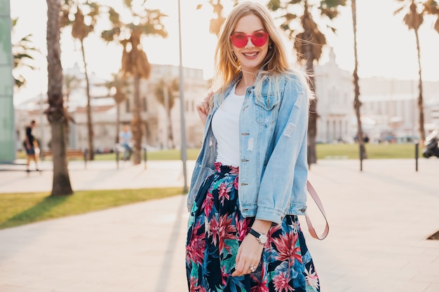 ピンクのサングラスを身に着けているスタイリッシュなプリントスカートとデニムの特大ジャケットで街の通りを歩いてかなり笑顔の女性、夏のスタイルのトレンド