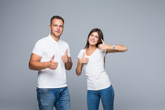 Pretty smiling young couple in casual clothing thumbs up isolated on grey