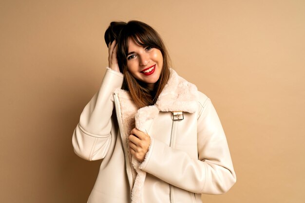 베이지색 배경에서 포즈를 취한 모피 겨울 코트를 입은 꽤 세련된 브루네트 여성, 밝은 유행 메이크업. 긍정적인 분위기, 블로거 복장.