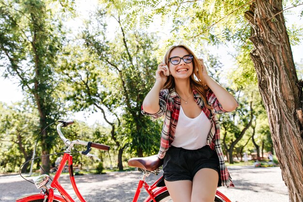 Pretty stylish female model enjoying morning in summer park. Outdoor portrait of laughing pleasant girl posing with bicycle.