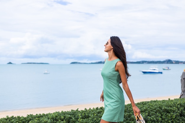 Pretty stylish happy woman in green summer dress with bag, wearing sunglasses on vacation, blue sea on background