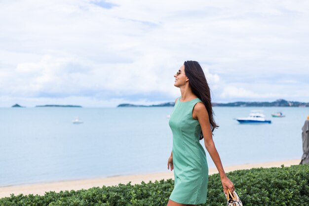 バッグと緑の夏のドレス、休暇でサングラス、背景に青い海を身に着けているかなりスタイリッシュな幸せな女性