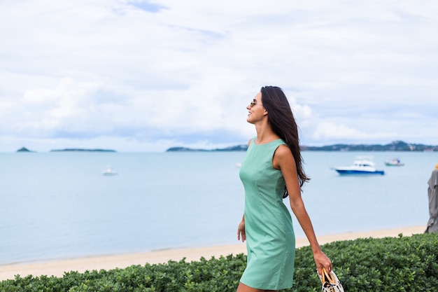 Довольно стильная счастливая женщина в зеленом летнем платье с сумкой, в темных очках на отдыхе, синее море на фоне