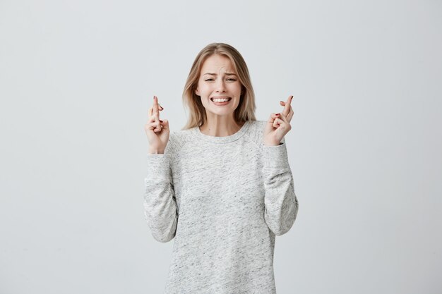 느슨한 스웨터에 염색 한 머리카락을 가진 매우 미신적 인 젊은 여성이 손가락을 교차시키고 중요한 행사 전에기도하고, 행운을 기원하며, 승리와 성공을 기원합니다. 희망을 느끼고 기적을 기다리는 여자