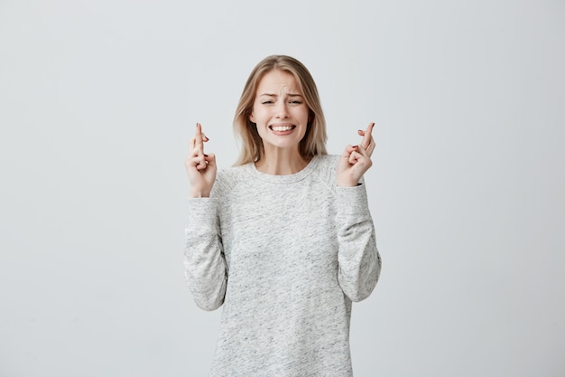 Pretty supertitious young woman with dyed hair in loose sweater crosses fingers, prays before important event, wishes luck, hopes for victory and success. Woman feeling hopeful, waiting for miracle