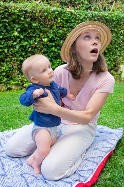 Pretty surprised mom sitting on plaid in park with open mouth and looking away. Cute baby girl in blue shirt standing with help of mother and looking aside