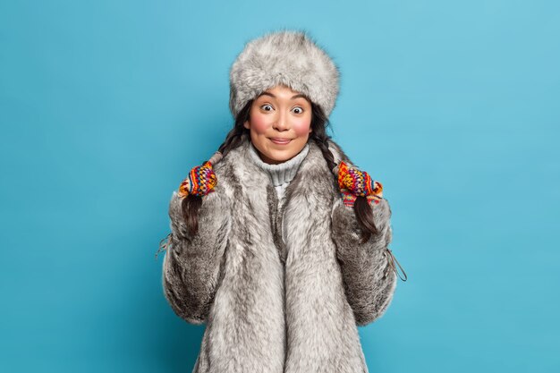 Pretty surprised young Asian woman holds pigtails dressed in outerwear isolated over blue wall. Eskimo female wears hat and coat lives in arctic place