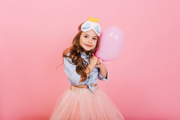Pretty sweet little girl with long brunette hair hugging balloon, looking to camera isolated on pink background. Beautiful joyful child having fun, celebrating birthday party, expressing positivity